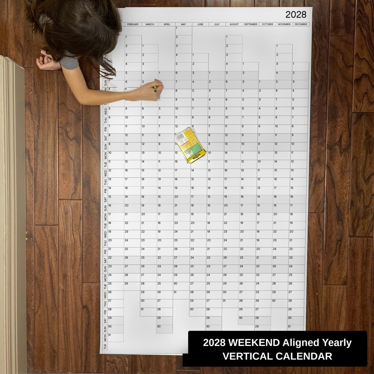 Child writing on the 2028 weekend-aligned yearly vertical giant paper wall calendar on a wood floor
