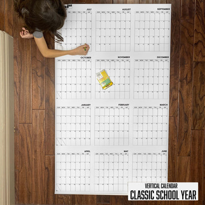 Child writing on the 2027-28 Classic School Year vertical giant paper wall calendar on a wood floor