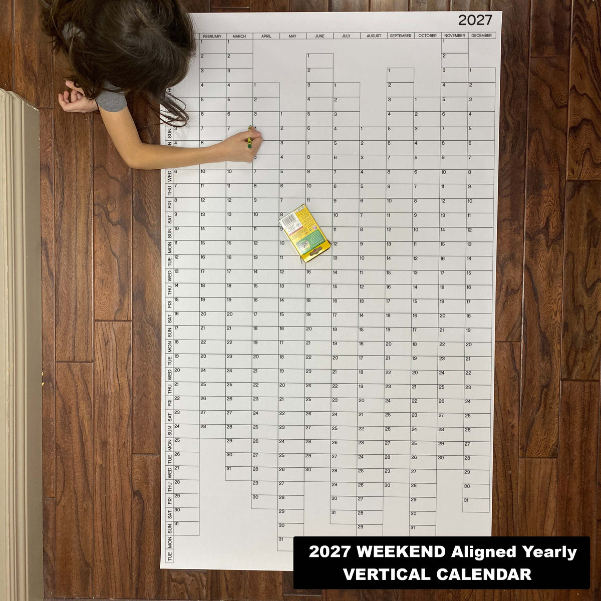 Child writing on the 2027 weekend-aligned yearly vertical giant paper wall calendar on a wood floor