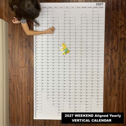 Child writing on the 2027 weekend-aligned yearly vertical giant paper wall calendar on a wood floor