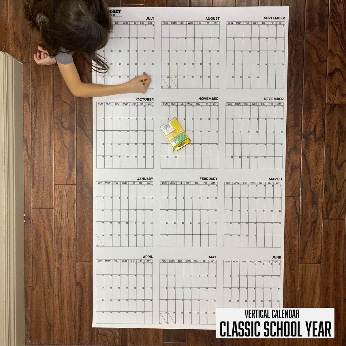 Child writing on the 2026-27 Classic School Year vertical giant paper wall calendar on a wood floor