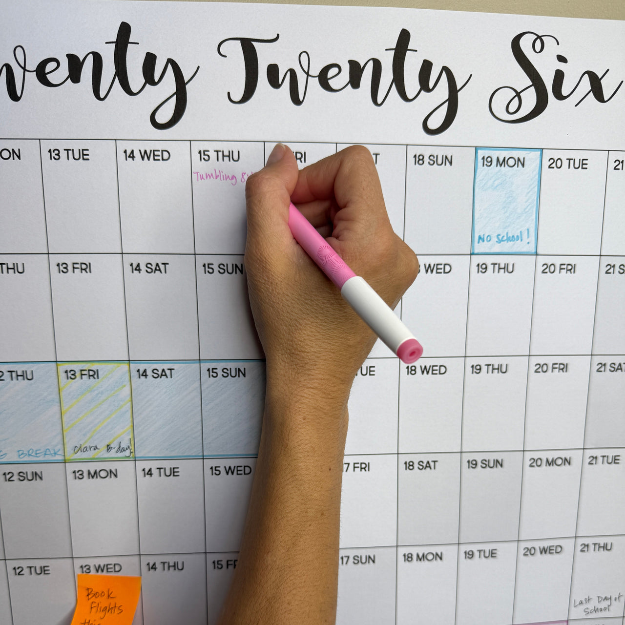 Hand holding a pink pen over a calendar with 'Twenty Twenty Six' text.